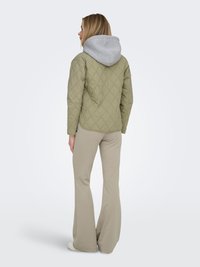 Kvinde med blondt hår iført en quiltet olivengrøn jakke med grå hætte, beige udvidede bukser, stående med ryggen mod en hvid baggrund.