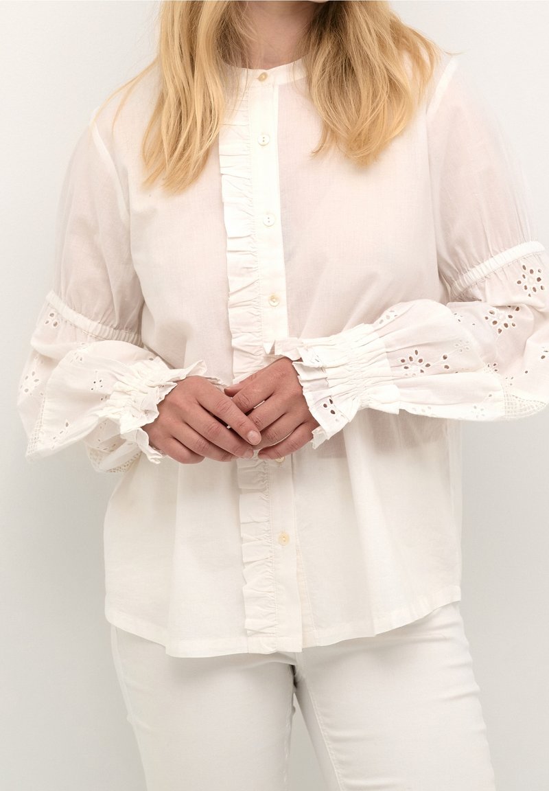 Weiße Baumwollbluse mit Rüschen an der Vorderseite und den Manschetten, verziert mit Lochstickerei. Lange Ärmel, lockere Passform und Knopfverschluss vorne.