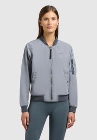 Femme portant une veste bomber gris clair avec fermeture éclair frontale, col, poignets et ourlet côtelés, et poche zippée sur la manche gauche, se tenant devant un fond gris.