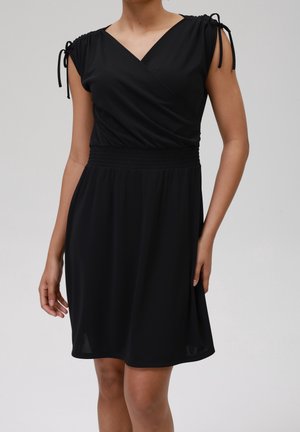 Kobieta w czarnej, bezrękawowej sukience do kolan z dekoltem w serek i marszczonymi detalami na ramionach związanymi na kokardę.