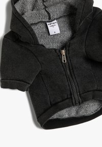 Sudadera negra con cremallera y forro de borreguillo gris. Presenta mangas cortas, puños de canalé y una textura suave. Etiqueta de talla visible en el interior.