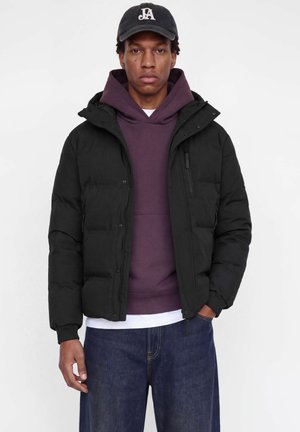 Un homme porte une doudoune noire par-dessus un sweat à capuche couleur prune, avec un jean foncé et une casquette noire à logo, sur un fond blanc uni.