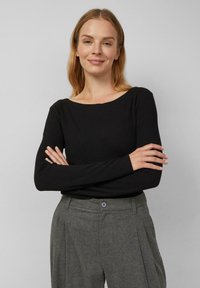 Kobieta z jasnobrązowymi włosami ubrana w czarną bluzkę z długim rękawem i szare spodnie z wysokim stanem, stojąca z założonymi rękami, na neutralnym tle.