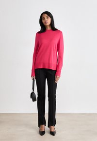 Roza pletena jopica s okroglim ovratnikom, v kombinaciji s črnimi krojenimi hlačami z razporki in črnimi špicastimi petkami. Majhna črna torbica.