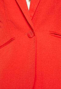 Blazer rouge avec un seul bouton rond, deux poches avant et une texture de tissu lisse. Coupe ajustée avec des revers nets.