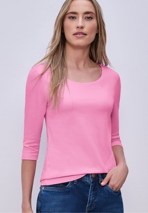 Donna con capelli biondi lunghi che indossa una maglia rosa a maniche 3/4 e jeans blu, in posa con una mano in tasca su uno sfondo semplice.
