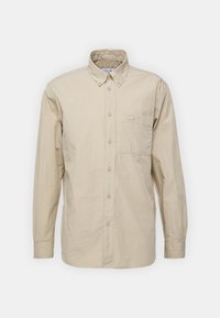 Camisa de botones de manga larga en color beige con cuello puntiagudo y un bolsillo en el pecho que presenta un pequeño logo bordado.