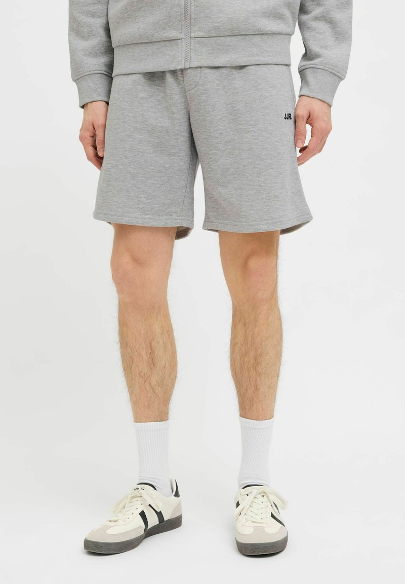Homme portant un short de sport gris avec un sweat à capuche assorti zippé, des chaussettes blanches et des baskets blanches à bandes noires, debout sur un fond blanc.
