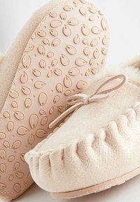 Chaussons mocassins beige doux avec un nœud, semelle en caoutchouc texturé anti-derrapante avec des motifs en forme de goutte, et doublure intérieure duveteuse visible.