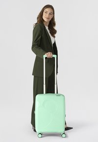 Valise rigide vert menthe à motif ondulé, équipée de roues doubles et d'une poignée télescopique, tenue par une personne en tenue formelle.