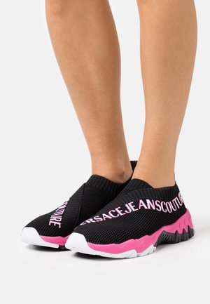 Baskets noires à enfiler avec semelles épaisses roses et blanches, et texte rose "VERSACE JEANS COUTURE", portées sur des jambes nues.
