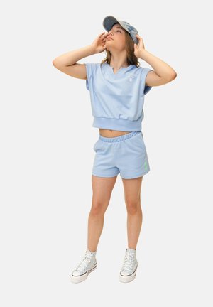 Young woman adjusting blue cap, wearing light blue sleeveless top and shorts, white high-top sneakers, standing against a plain background.