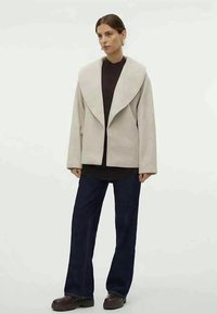 Mulher a usar um casaco oversized bege com grande colarinho, blusa preta, calças largas escuras e sapatos castanhos chunky, de pé contra um fundo liso.