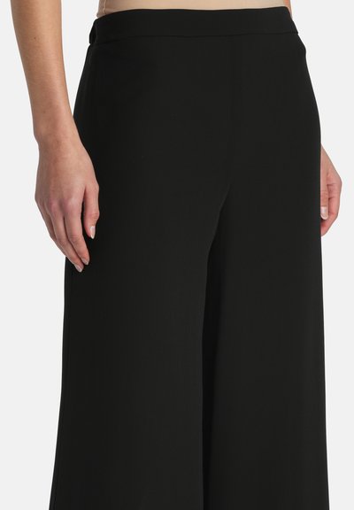 Femme portant un pantalon noir taille haute à jambes larges avec une ceinture plate, les mains détendues le long du corps, sur un fond clair uni.