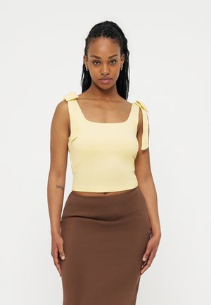 Femme aux cheveux tressés portant un haut jaune sans manches avec des bretelles nouées aux épaules et une jupe marron taille haute, se tenant devant un fond blanc.