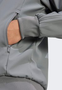 Szara kurtka bomberka z gładką teksturą, ściągaczami przy mankietach i boczną kieszenią na zamek. Tkanina ma lekki połysk.