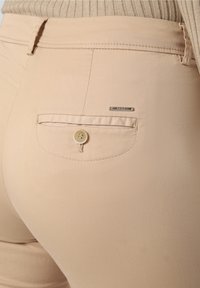 Zbliżenie na beżową kieszeń spodni z guzikiem i małą metalową zawieszką z napisem "BRAX" na prążkowanym beżowym tle.