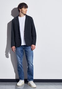 Svart kavaj med två knappar och fickor, bärs över en vit t-shirt. Kombineras med ljusblå jeans och vita sneakers mot en enkel bakgrund.