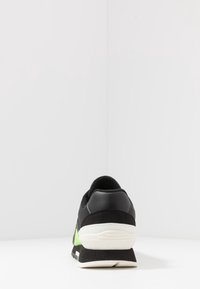 Černé tenisky s síťovaným svrškem, zelenými akcenty, polstrovaným límcem a silnou bílou podrážkou. Vyznačují se hladkým povrchem a texturovanou boční panelem.