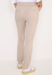 Beige sportovní kalhoty s hladkou texturou, elastickým pasem a dvěma zadními kapsami. Nosené s bílými teniskami s modrými akcenty.