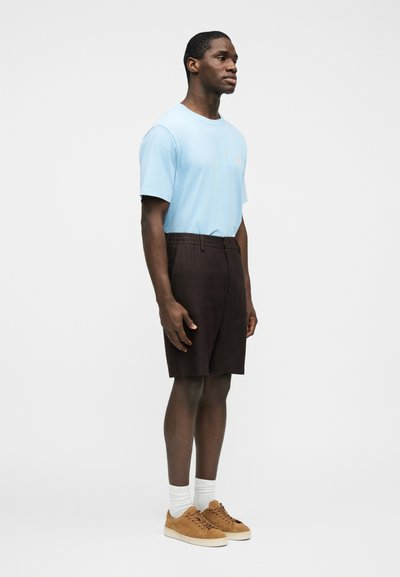 Homme portant un t-shirt bleu clair, un short marron foncé, des chaussettes blanches et des baskets en daim beige, debout devant un fond blanc.