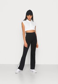Blusa branca cropped com mangas curtas, combinada com calças pretas largas com riscas brancas nas laterais e um boné preto. Sapatilhas brancas completam o visual.