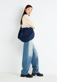 Sac en tissu bleu marine au design décontracté, associé à un pantalon en jean large bleu clair et des bottes à talons épais noires. Un pull neutre est porté.