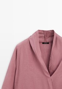 Camicia di lino color malva con scollo a V profondo e collo a scialle, che mostra la metà superiore e l'etichetta del marchio all'interno del collo.