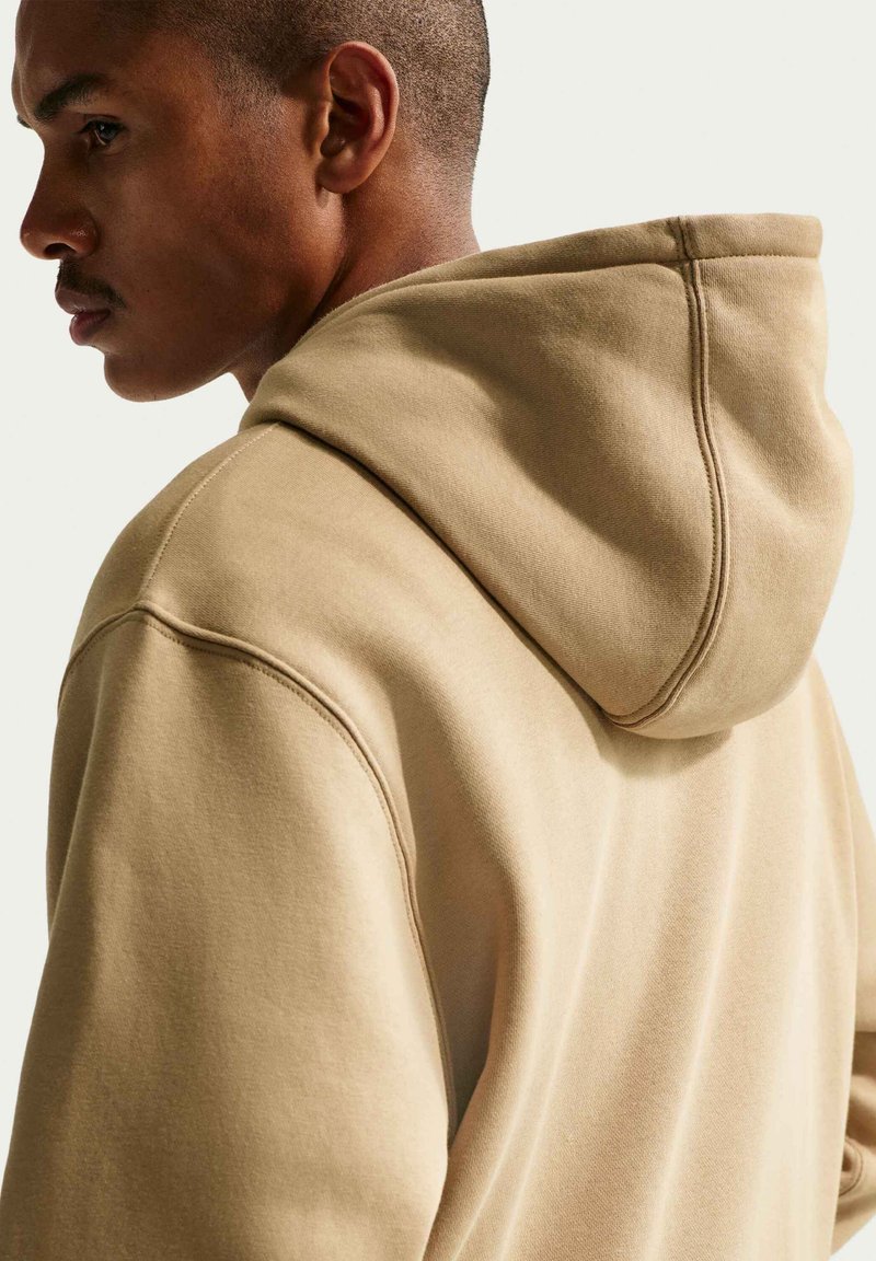 Mann mit kurzen Haaren, der einen beigen Hoodie trägt, von der Seite und von hinten vor einem einfarbigen hellen Hintergrund gezeigt.