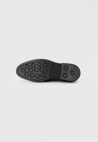 Suela de zapato de goma negra con un patrón de tracción circular, punta redondeada y tacón ligeramente elevado. Superficie texturizada para mejor agarre.