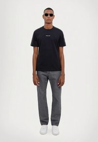 Melna kokvilnas T-krekls ar baltu logotipu, īsām piedurknēm un kontrastējošām svītrām uz pleciem. Kopā ar pelēkām džinsa biksēm un baltiem sporta apaviem.