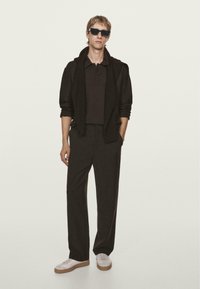 Polo nero sotto un maglione zip-up abbinato, abbinato a pantaloni neri larghi e sneakers bianche. Il modello indossa occhiali da sole scuri.