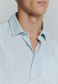 Chemise à boutons en bleu clair avec un petit motif géométrique, dotée d'un col pointu et d'une poche poitrine. Le tissu semble lisse et léger.
