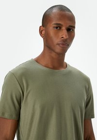 Camiseta de algodón verde oliva con cuello redondo, mangas cortas y sutil textura acanalada. Diseño minimalista, sin patrones ni acentos visibles.