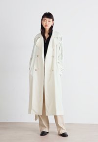 Trench-coat beige clair avec un design à double boutonnage, grands revers et poches latérales. Associé à un top noir et un pantalon beige.