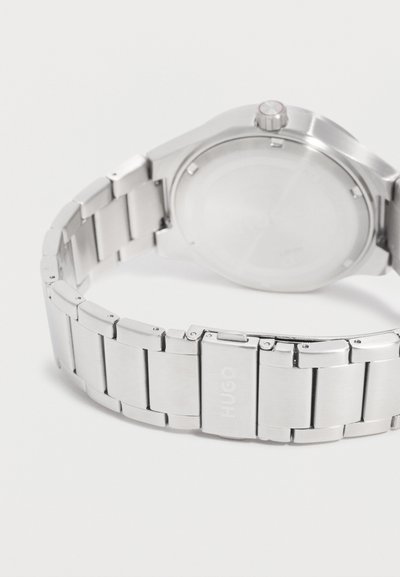 Bracelet de montre en métal argenté avec un fermoir gravé "HUGO", fixé à l'arrière d'un boîtier de montre rond avec une couronne sur le côté droit.