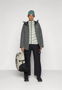 Siva vodoodporna jakna s svetlo sivo puhovino notri, črne cargo hlače, zelena pletena kapa in bež-črn nahrbtnik. Gladke teksture.