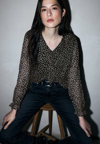 Bruine bloemige blouse met V-hals en gerimpelde mouwen, gecombineerd met zwarte high-waisted jeans. Model zittend op een houten kruk.