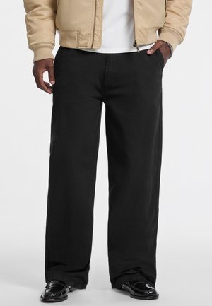 Black wide-leg trousers made from a durable fabric. Features a flat front, side pockets, and hemmed cuffs. Paired with glossy shoes.