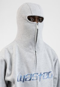 Grå fleece hoodie med høj krave, lynlås foran og øjeåbninger. Har et blåt logo på brystet, der viser et unikt design.