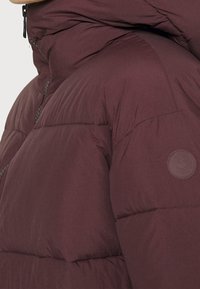 Bordeauxfarbene Pufferjacke mit hohem Kragen, strukturierten Oberflächen und einem runden Logopatch am linken Ärmel. Verfügt über einen Reißverschluss vorne und eine detaillierte Steppnaht.