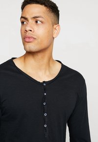 Camisa negra de manga larga con cuello redondeado, cierre de botones con botones azules, y una tela suave y texturizada.