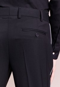 Svarta skräddarsydda byxor med en texturerad yta, utrustade med en sidficka med knapp och vit piping längs sömmen.