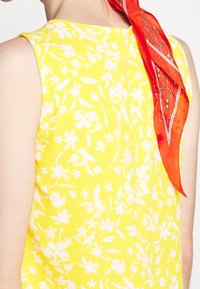 Ärmlös gul blus med ett vitt blommönster, texturerat tyg och en halsduk i rött med ett geometriskt mönster.