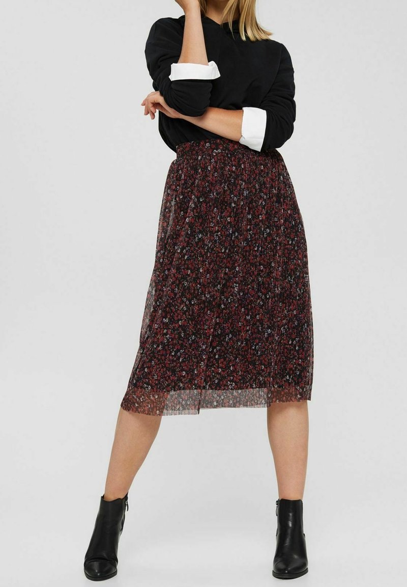 Femme portant un haut noir à manches longues avec des poignets blancs, une jupe midi plissée fleurie rouge et noire, et des bottines noires, debout, bras croisés.