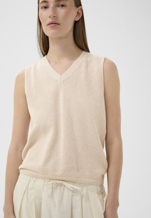 Femme portant un gilet en tricot beige sans manches avec un col en V et un pantalon plissé crème clair taille haute avec cordon de serrage.