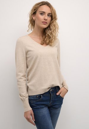 Maglione beige a V, realizzato in materiale morbido, con maniche lunghe e orlo a coste. Indossato con jeans aderenti blu.