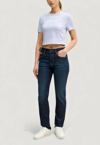 Biały krótki t-shirt z nadrukowanym logo, zestawiony z ciemnoniebieskimi prostymi dżinsami i białymi trampkami. Dżins lekko błyszczy.