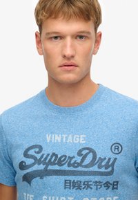 Niebieski, melanżowy bawełniany t-shirt z logo "SuperDry" w ciemnoszarym kolorze, okrągłym dekoltem, krótkimi rękawami i luźnym krojem.