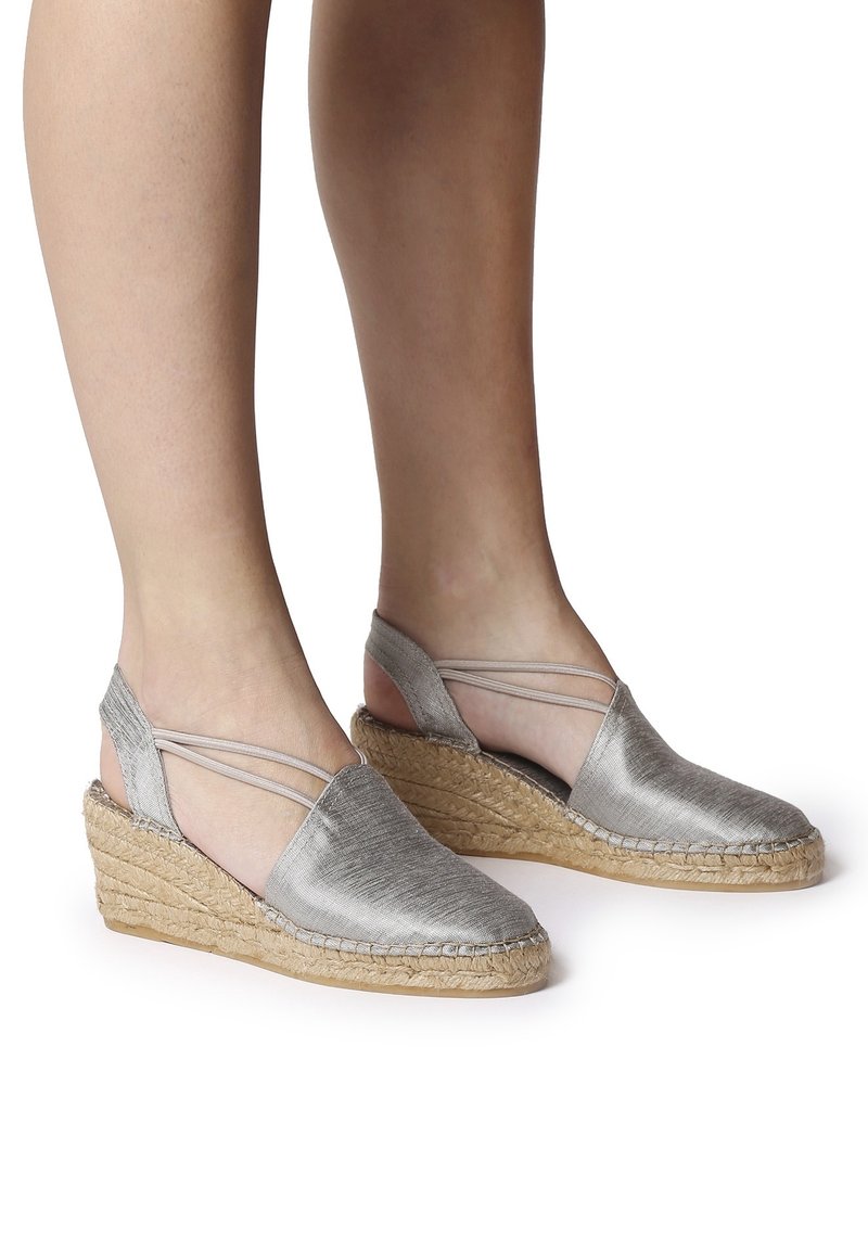Pieds portant des espadrilles compensées argentées à bride arrière avec semelles plateforme en jute tressée sur fond blanc.